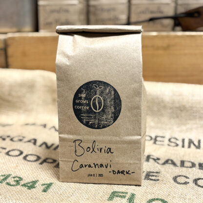 Product Shot: Recycled kraft bag filled with fresh roasted coffee, labelled "Bolivia Caranavi Dark", branded with a She Grows Coffee stamp, and stamped with a roast date.