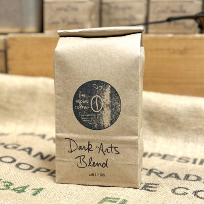 Product Shot: Recycled kraft bag filled with fresh roasted coffee, labelled "Dark Arts Blend", branded with a She Grows Coffee stamp, and stamped with a roast date.