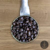 Close-up shot of Dark Arts Blend dark roasted coffee beans in a handmade scoop with a wooden cutting board background.
