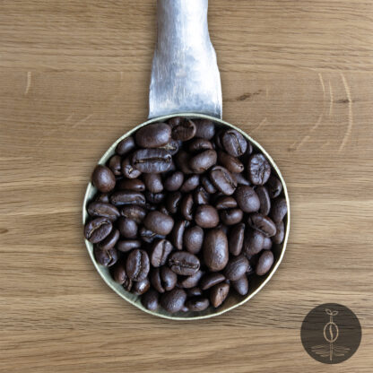 Close-up shot of Dark Arts Blend dark roasted coffee beans in a handmade scoop with a wooden cutting board background.